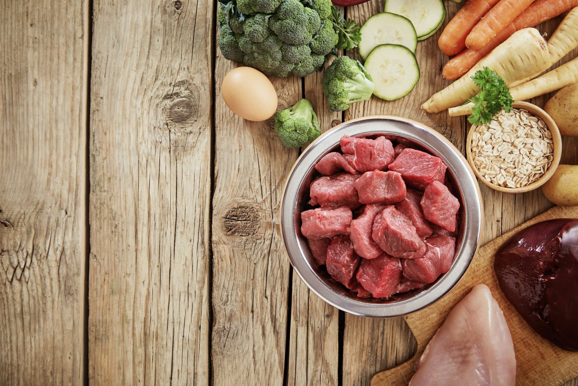 Fleisch und Gemüse als Zutaten für ein vitaminreiches Katzenfutter um einen Napf mit Frischfleisch.
