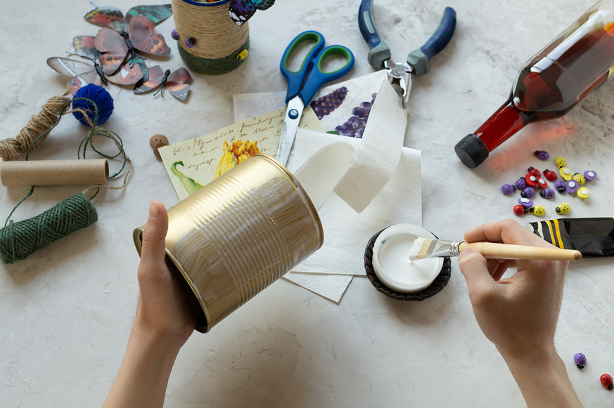 Zwei Hände sind über einem Basteltisch mit Scheren, Garn, Papier und Dekoelementen zu sehen, wie sie eine Blechdose mit weißer Farbe bemalen. 
